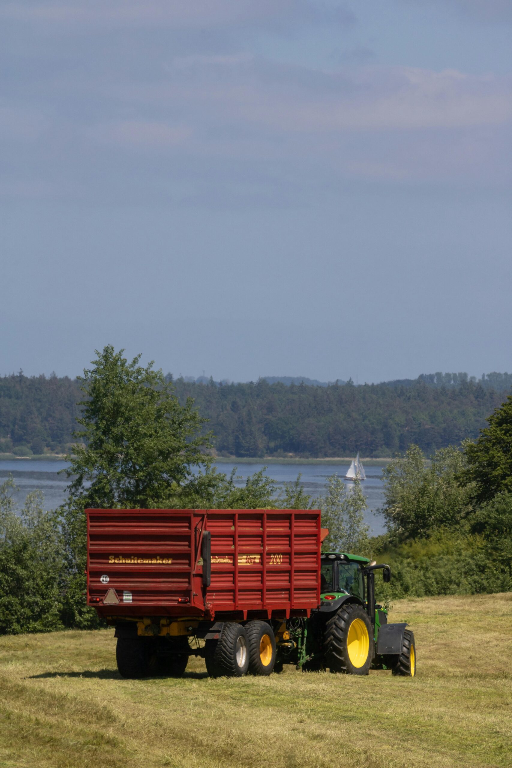 Tractor with red trailer in rural field by a lake with sailboat.